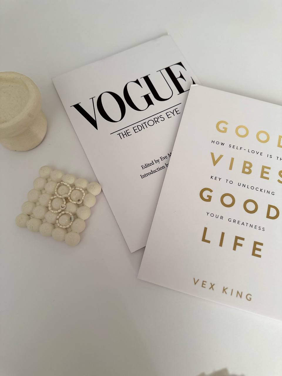 Two books, one titled 'Vogue' and the other 'Good Vibes Key to Unlocking Your Greatestness', on a white surface with a small decorative item.